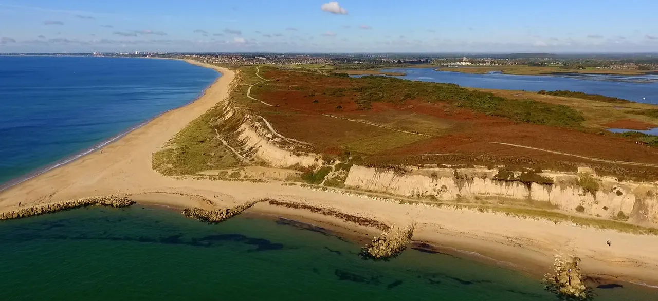 Aerial view of a sandy beach with white chalk cliffs, blue sea, and coastal town in the distance.