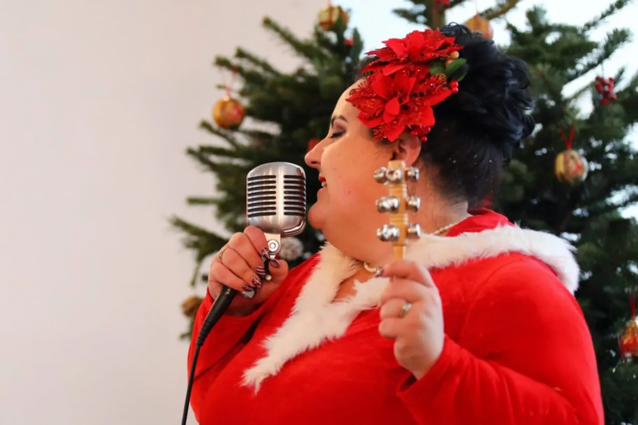 Woman in red Father Christmas outfit with red flower headpiece singing into vintage microphone in front of decorated Christmas tree.