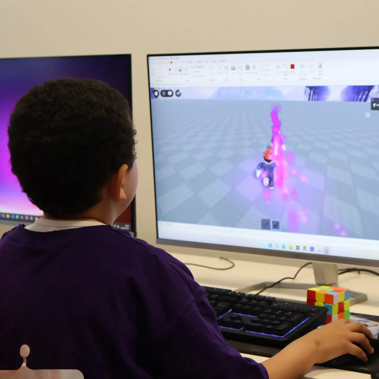 Child in purple shirt playing a block-building video game on screen at a computer station with keyboard.