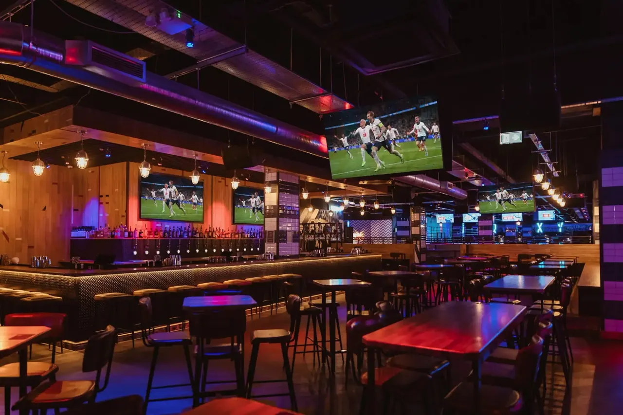 Modern sports bar interior with multiple screens showing a football match, neon-lit bar counter, and dining tables in red and blue lighting.