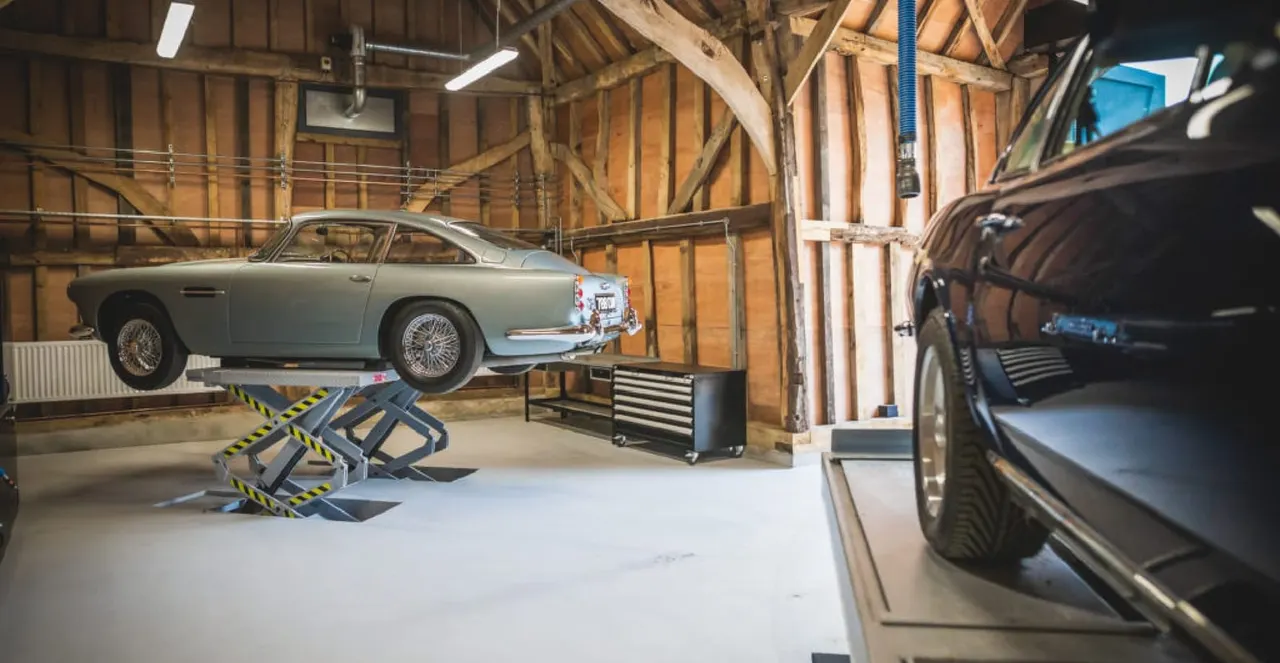 Classic sports car on a hydraulic lift in a timber-framed garage workshop with other vintage vehicles.