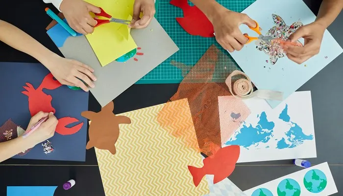 Multiple children's hands working on craft projects with coloured paper, scissors, and decorative materials at a table.