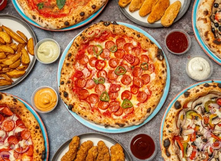 Overhead view of multiple pizzas with various toppings, potato wedges, and sauce bowls on a grey surface.