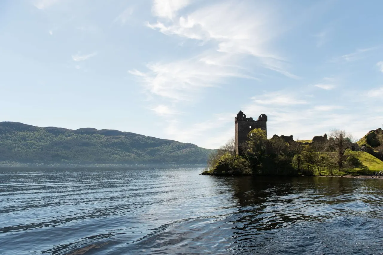 Ruined castle on a peninsula in a large loch, surrounded by water and forested hills under blue sky.