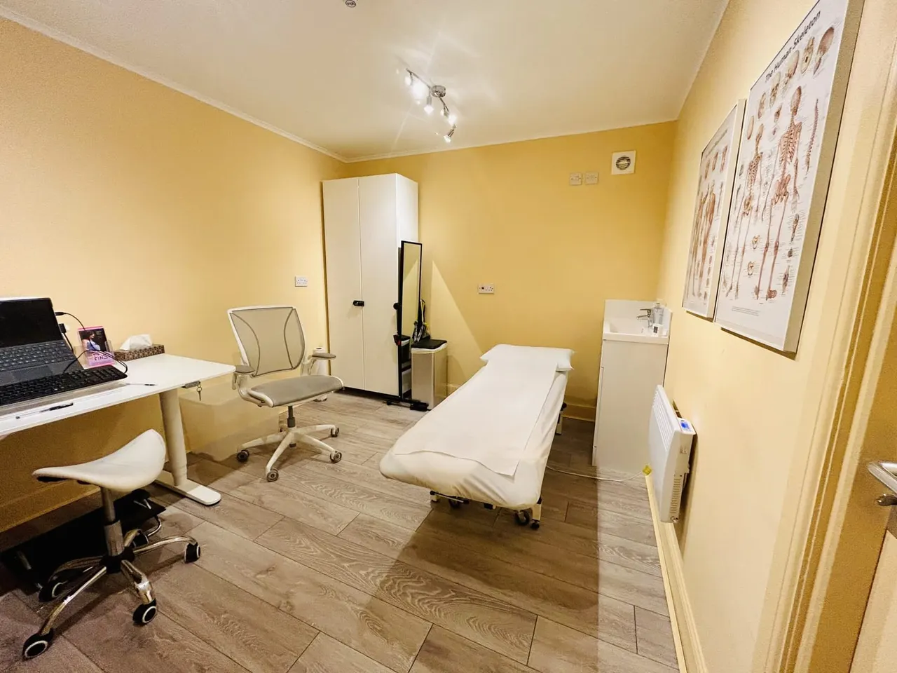 A bright yellow therapy or treatment room with a white massage couch, desk with computer, office chair and anatomical charts on the walls.
