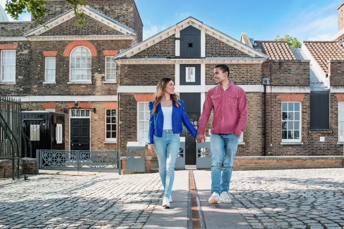 Two people walk hand in hand along the Prime Meridian line on a paved courtyard in front of historic brick buildings.