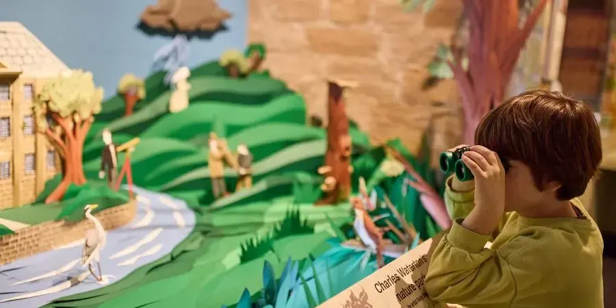 A young child using binoculars to view a colourful diorama display featuring a landscape with buildings, fields and trees.