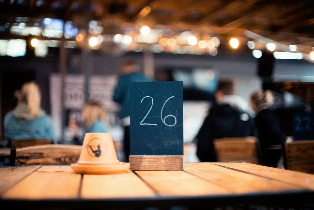 Table number 26 with wooden base and ceramic holder on dining table, diners and warm lighting in soft-focus background.