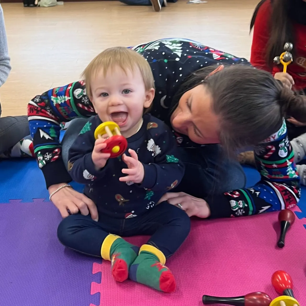 A laughing toddler sits on a padded play mat holding a toy while an adult leans in close beside them during a Baby College class session.