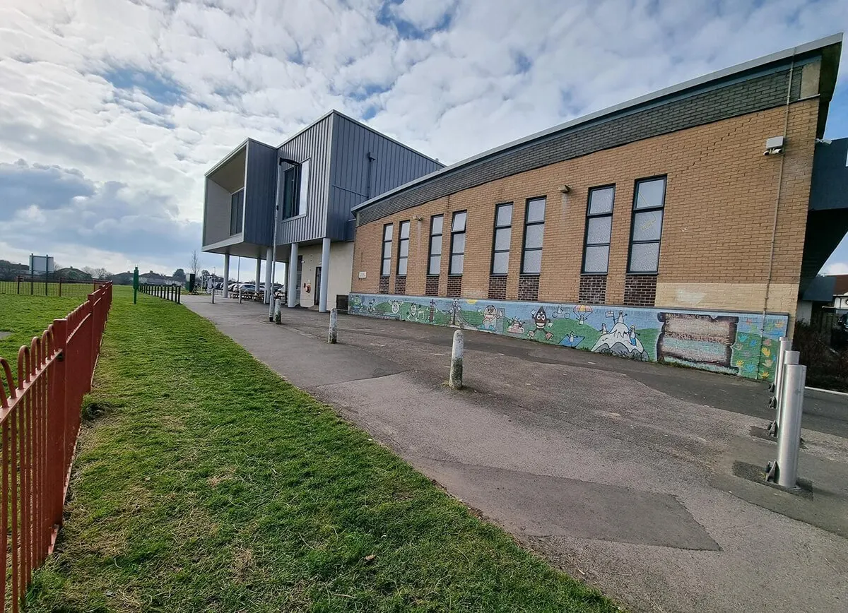 Modern brick and metal leisure building with colourful mural, surrounded by open grass and paved areas under cloudy sky.