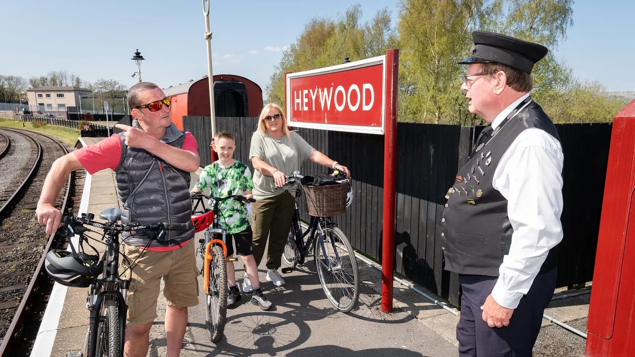 A family with bicycles meets a uniformed railway staff member at Heywood station platform, beside a red and black train carriage.