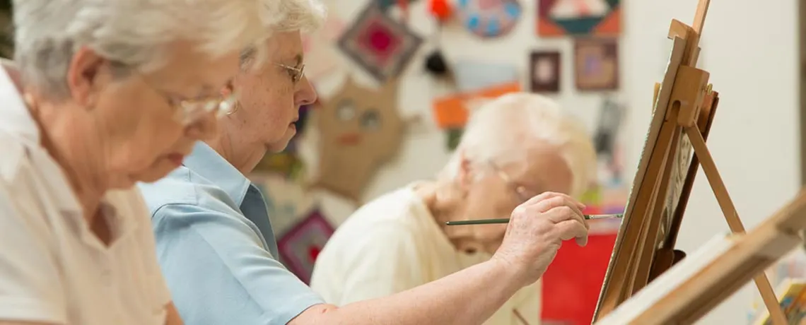 Older adults painting at easels in an art studio with artwork displayed on the walls behind them.