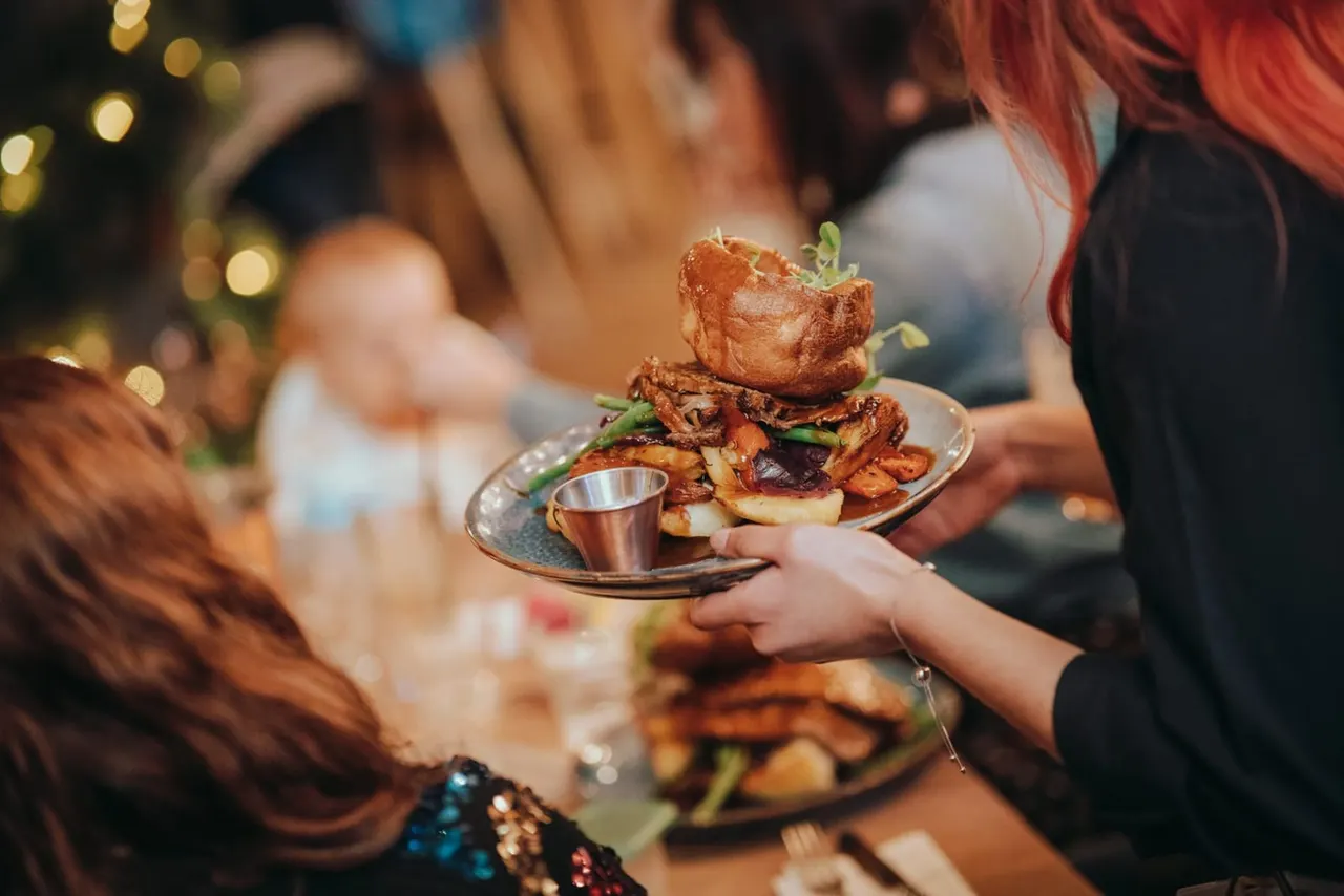 Person holding a plate of roasted meat stacked with vegetables and a dipping sauce at a dining table with other diners in the background.