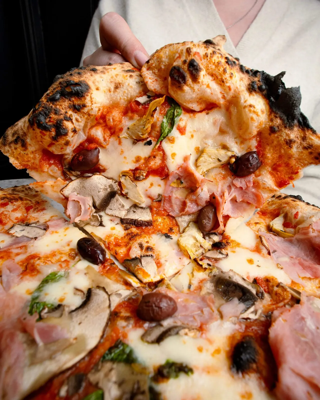 A person holds a slice of wood-fired pizza topped with cheese, ham, olives, artichokes and herbs.