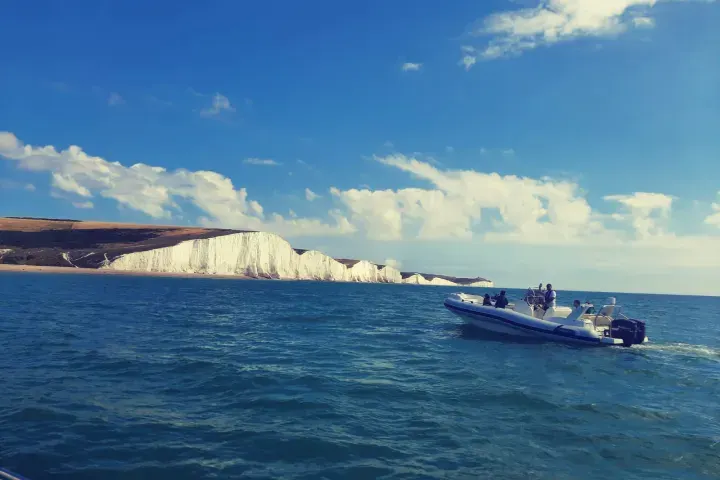 Blue speedboat with passengers travelling past white chalk cliffs on a calm sea under blue sky.