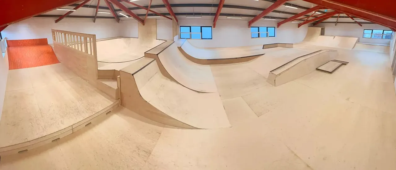 Interior of indoor skatepark with wooden ramps, curves and obstacles under red roof beams and skylights.