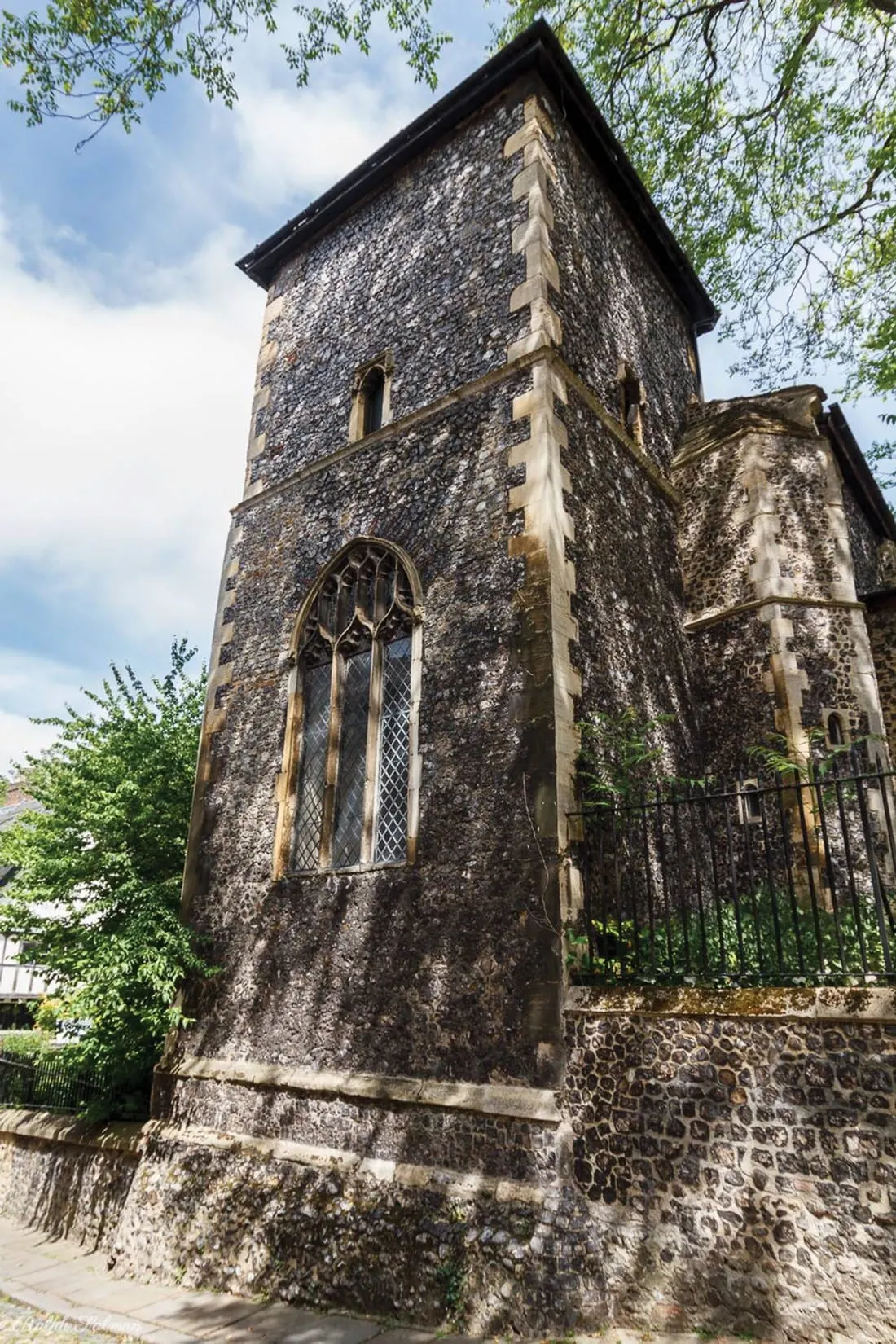 Historic stone church tower with Gothic window and weathered grey walls, surrounded by green foliage.