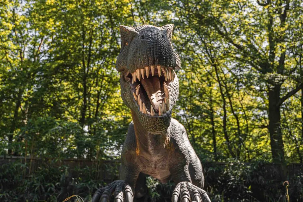 Large dinosaur sculpture with open mouth displaying teeth, set amongst green woodland foliage.