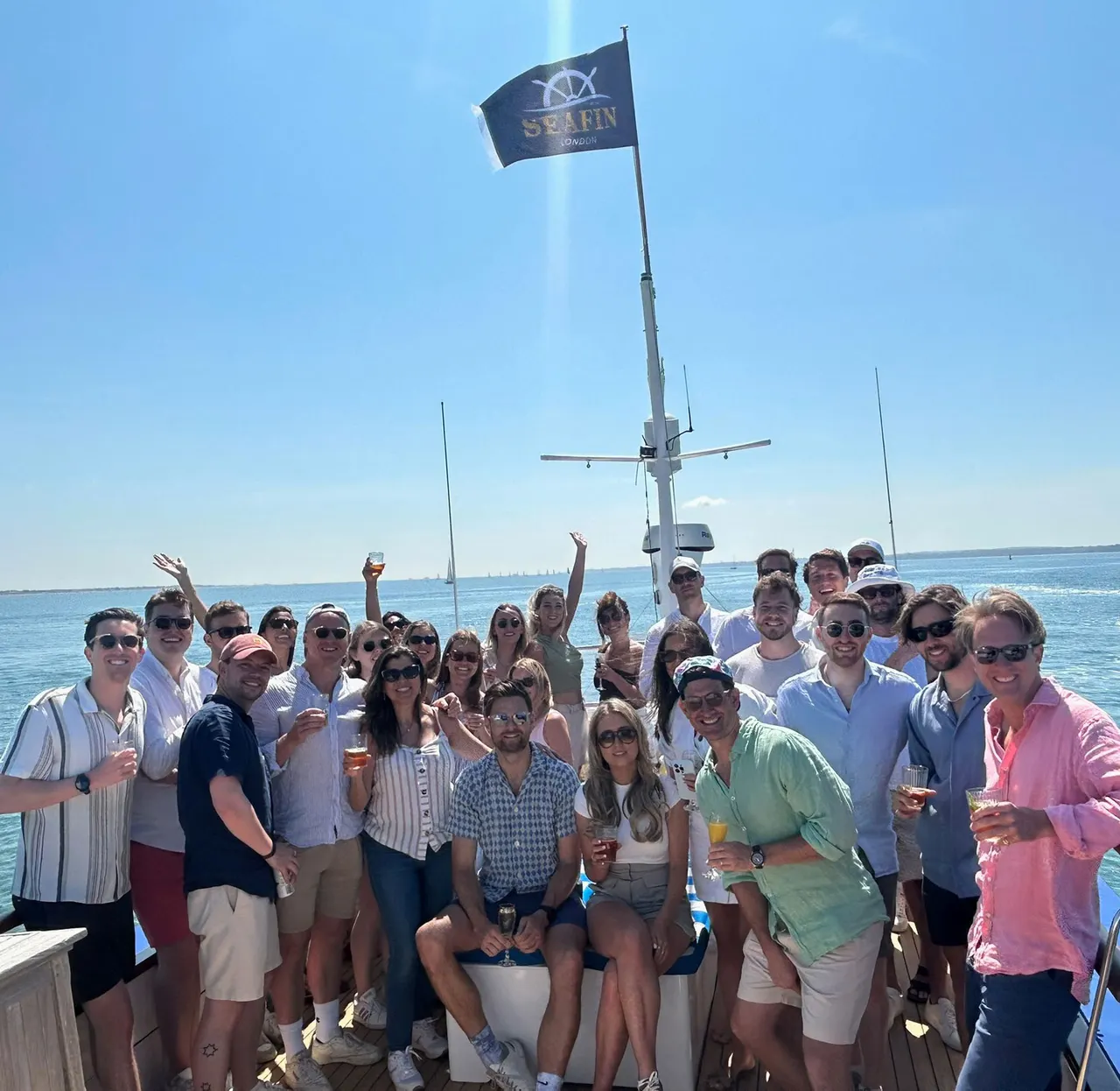 Large group of people gathered on a boat deck with drinks, clear blue sea and sky behind them.