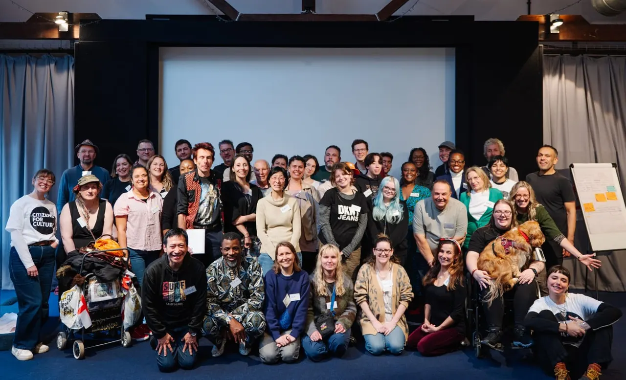 A diverse group of around fifty people posing together on stage for a formal group photograph at an indoor venue.