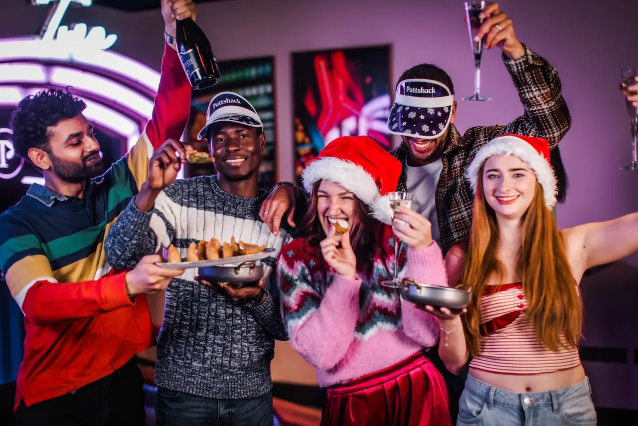 Group of five people celebrating at a golf venue, some wearing Santa hats, holding champagne and food under neon lighting.