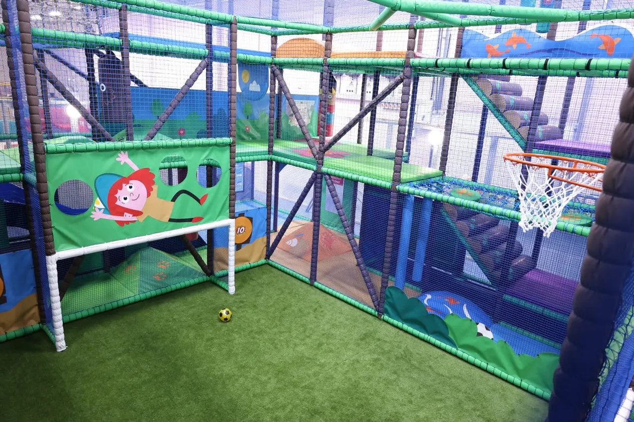 Inside a soft play area with colourful padded structures, a small football goal, basketball hoop and themed wall panels featuring cartoon characters.