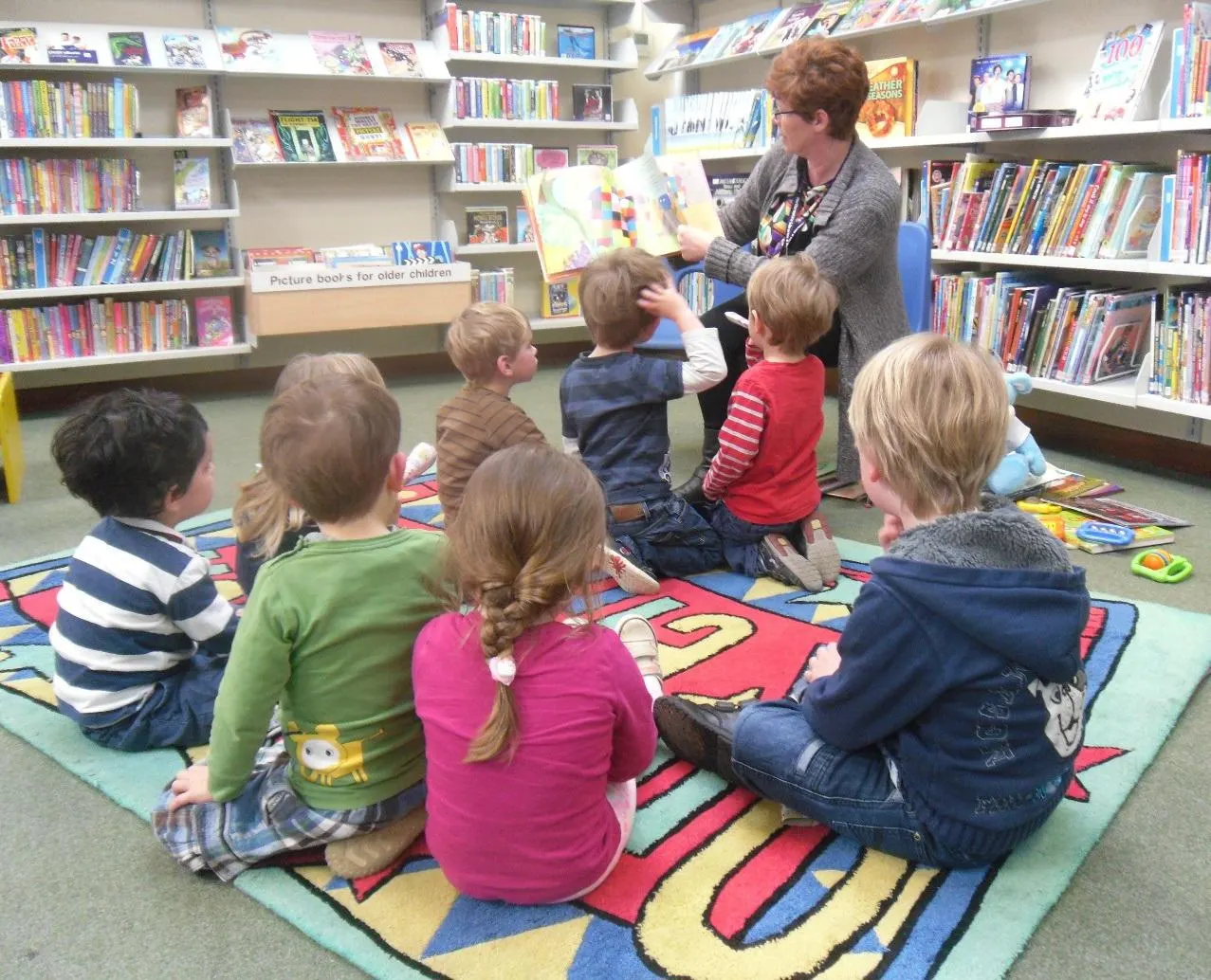 Adult librarian showing a picture book to a group of young children sitting on a coloured mat in a library surrounded by bookshelves.