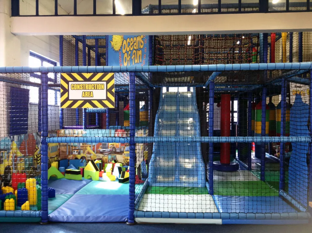 Indoor soft play area with blue netting, slides, climbing structures and colourful foam blocks for children.