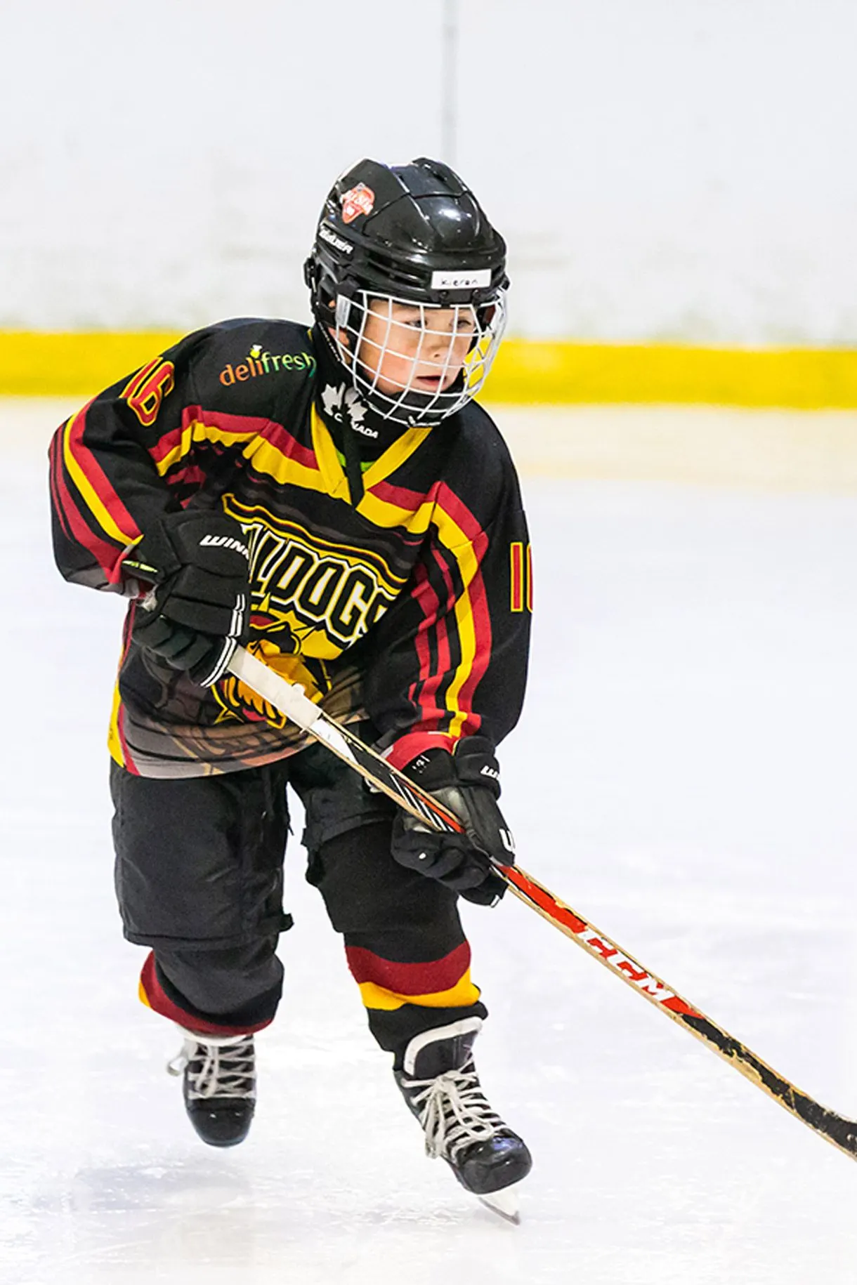 Young ice hockey player in black, red and yellow team kit holding a stick on the ice.