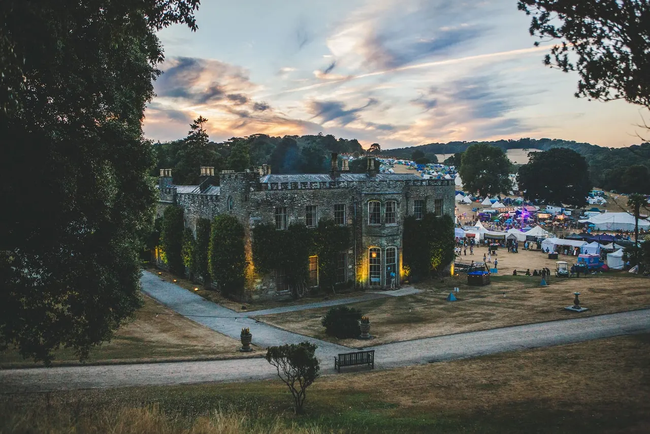 Stone mansion with illuminated grounds overlooking a festival site with colourful tents and crowds beside a lake at dusk.