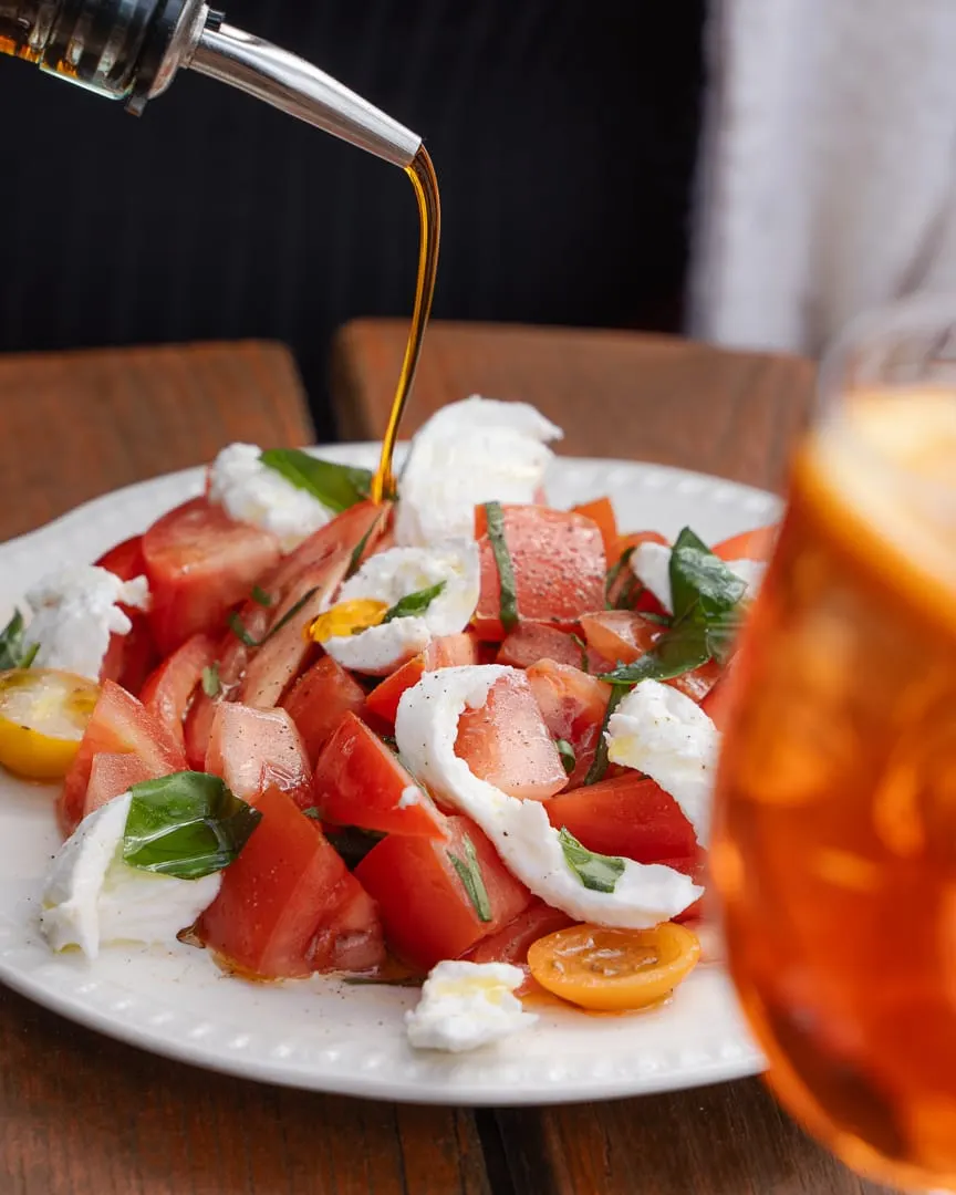 Olive oil being drizzled over a caprese salad with tomatoes, mozzarella and basil on a white plate.