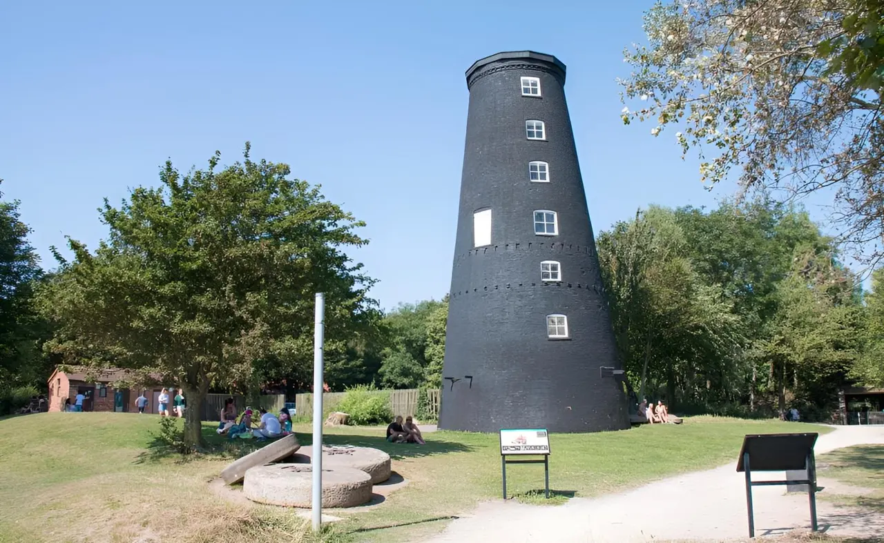 A historic brick windmill surrounded by green grass and trees, with visitors relaxing on the lawn beneath dappled shade.