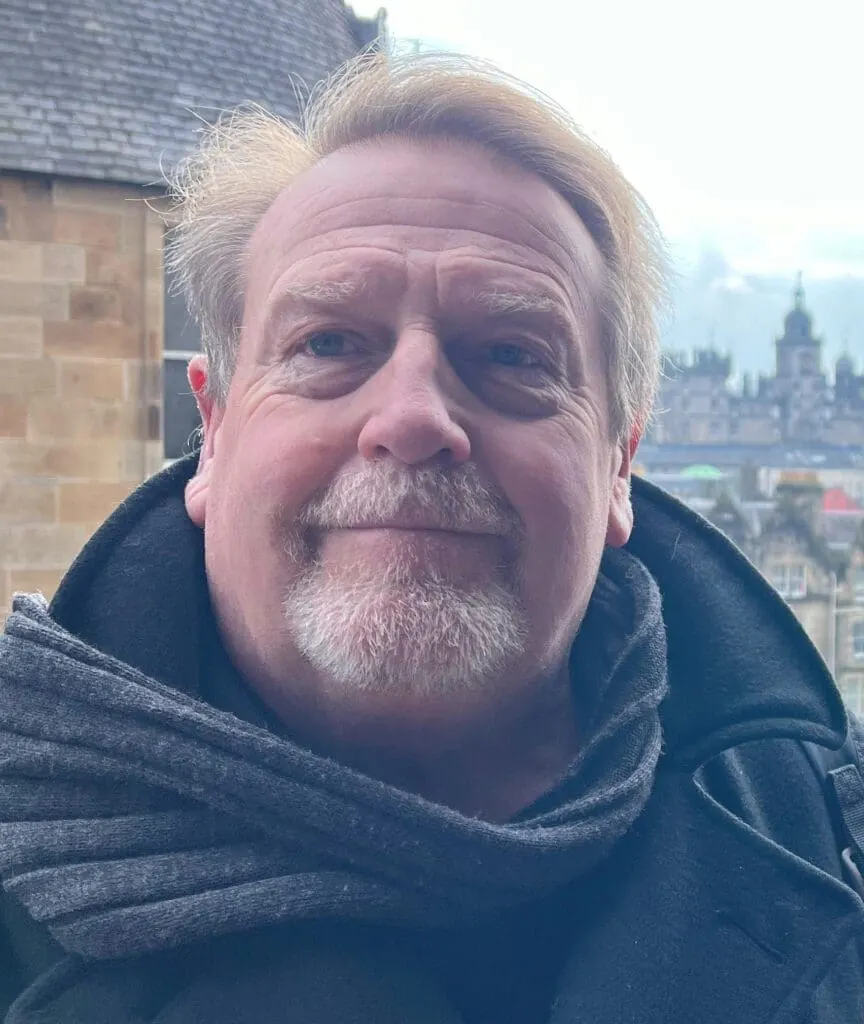 Man with beard posing for selfie with Edinburgh cityscape and historic buildings in background.