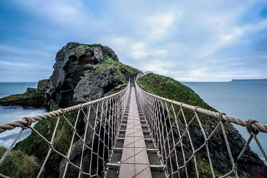 Suspension bridge spanning between two moss-covered rocky islands with sea visible below under a blue cloudy sky.