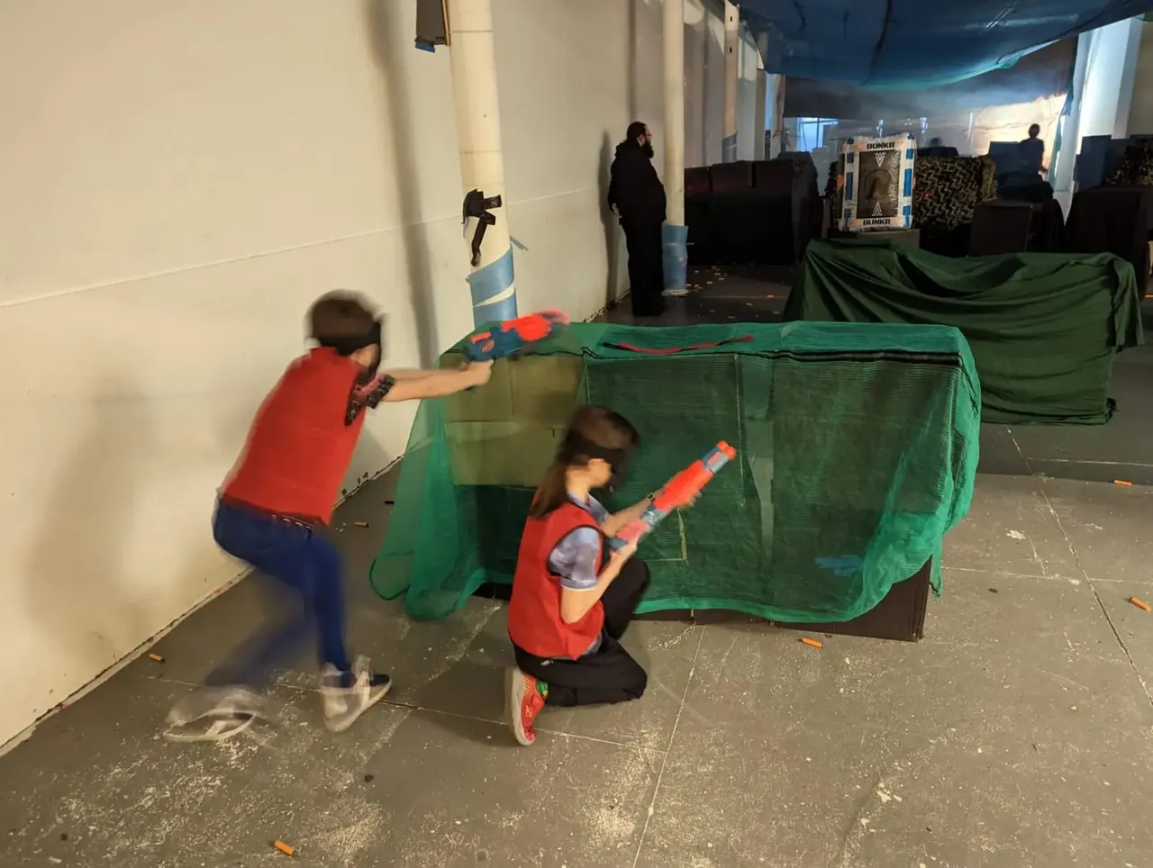 Children playing with foam dart blasters near green mesh barriers in an indoor arena.