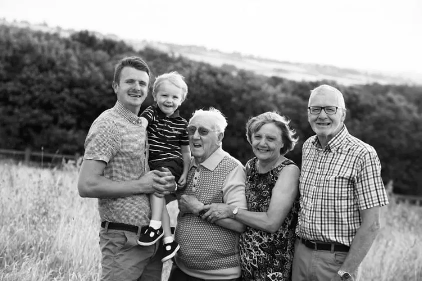 Black and white photograph of a family group of five people, including a young child, standing together in a rural field with woodland in the background.