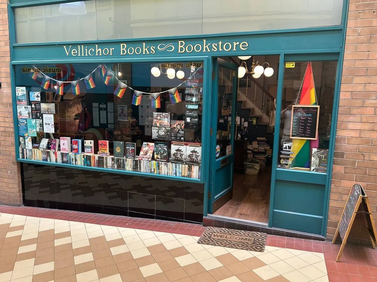 Turquoise-fronted independent bookstore with rainbow bunting in window display, books visible on shelves, rainbow flag decoration inside.