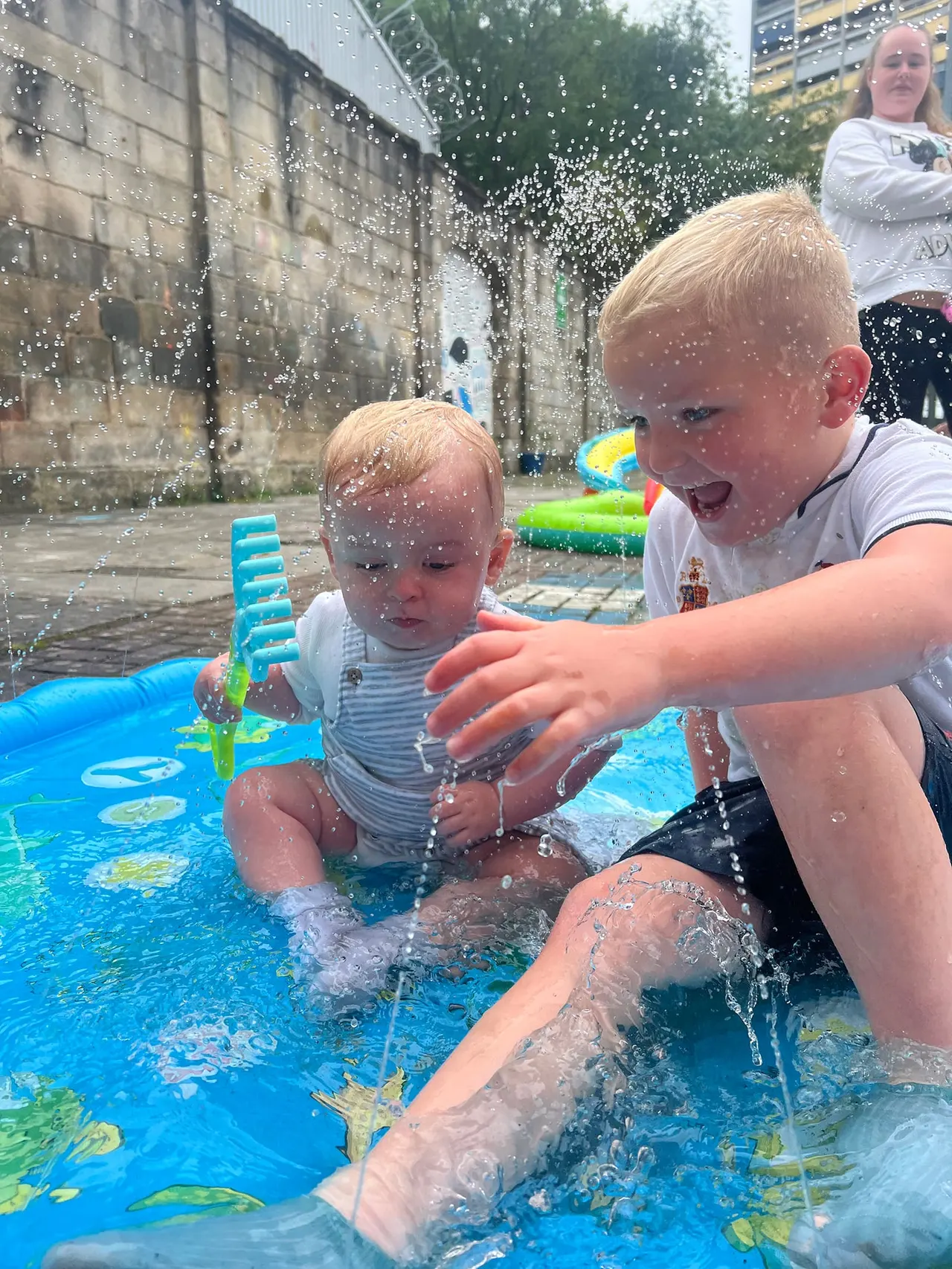 Two young children playing in a shallow paddling pool with water toys, supervised by an adult in an urban courtyard setting.