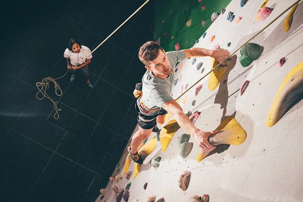 Man climbing an indoor climbing wall with coloured holds, belayed by an instructor below.