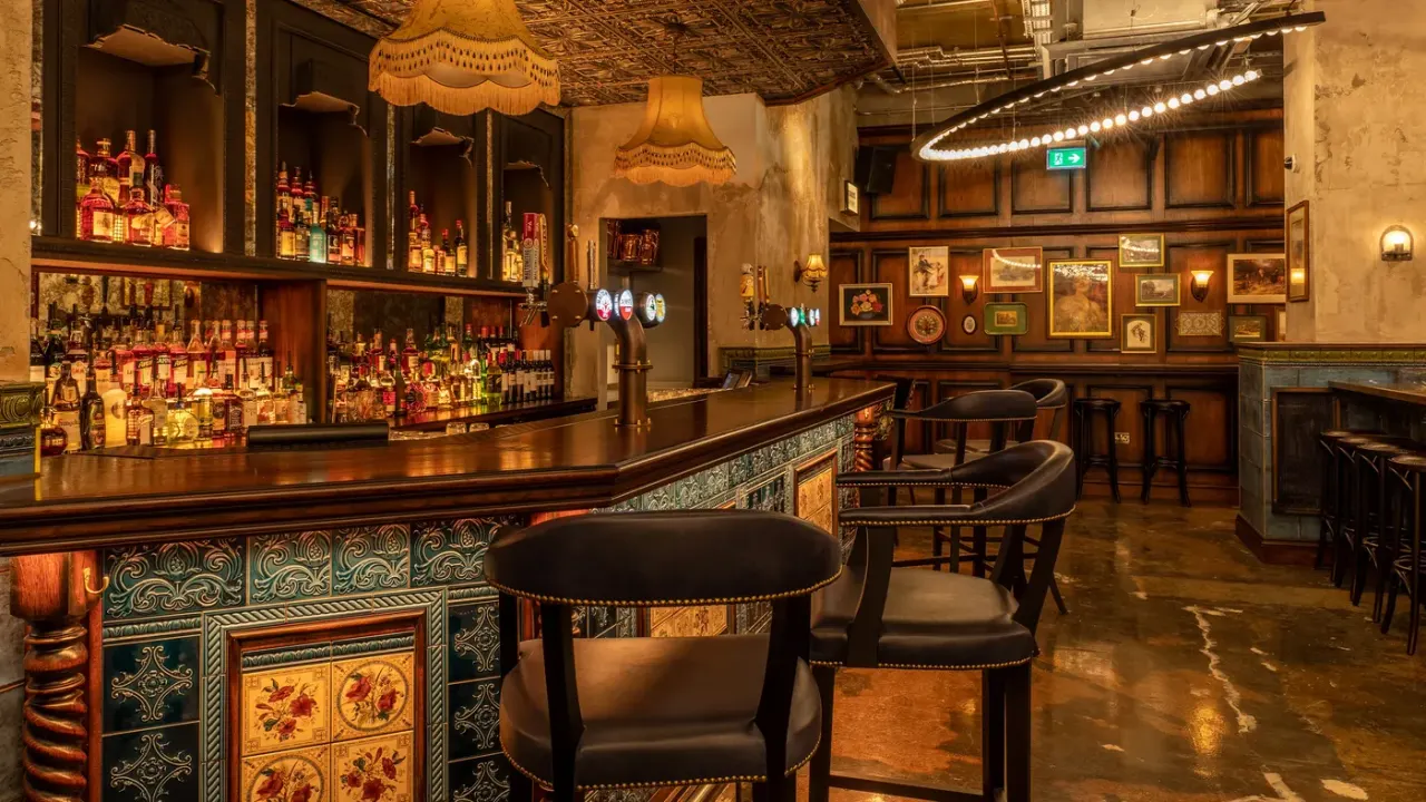 An upscale bar interior with a long wooden counter, shelves of spirits, decorative pendant lights, framed artwork, and leather seating areas.