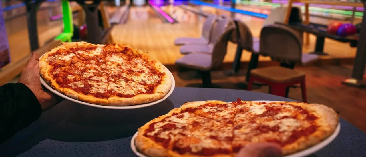 Two pepperoni pizzas on a table with bowling lanes and seating visible in the background under coloured lighting.