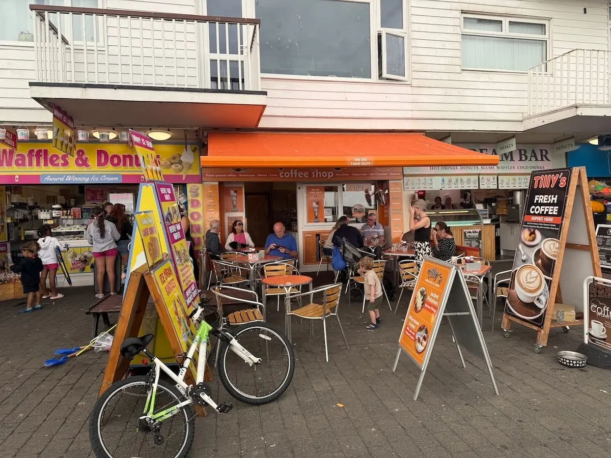 Busy seafront promenade with cafés, visitors seated at outdoor tables, and bicycles parked nearby.