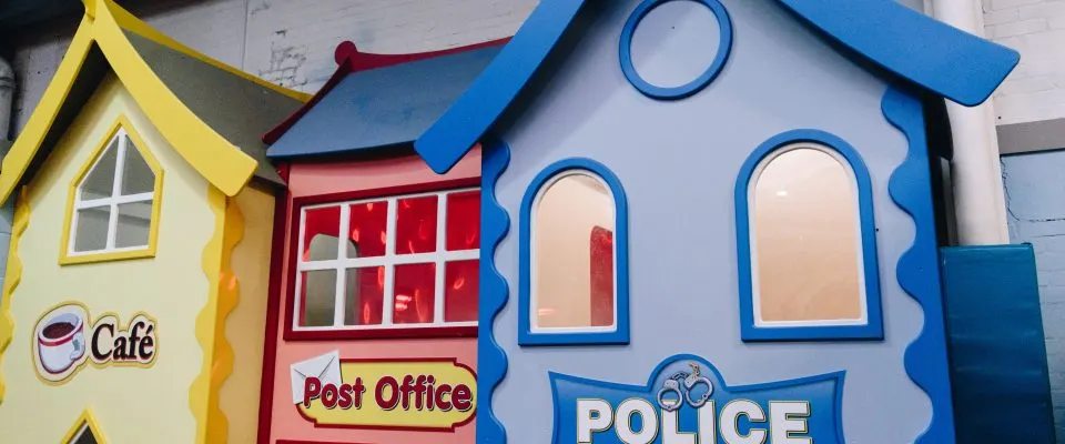 Colourful play structures designed as fairy-tale buildings labelled Café, Post Office and Police in bright yellow, red and blue.