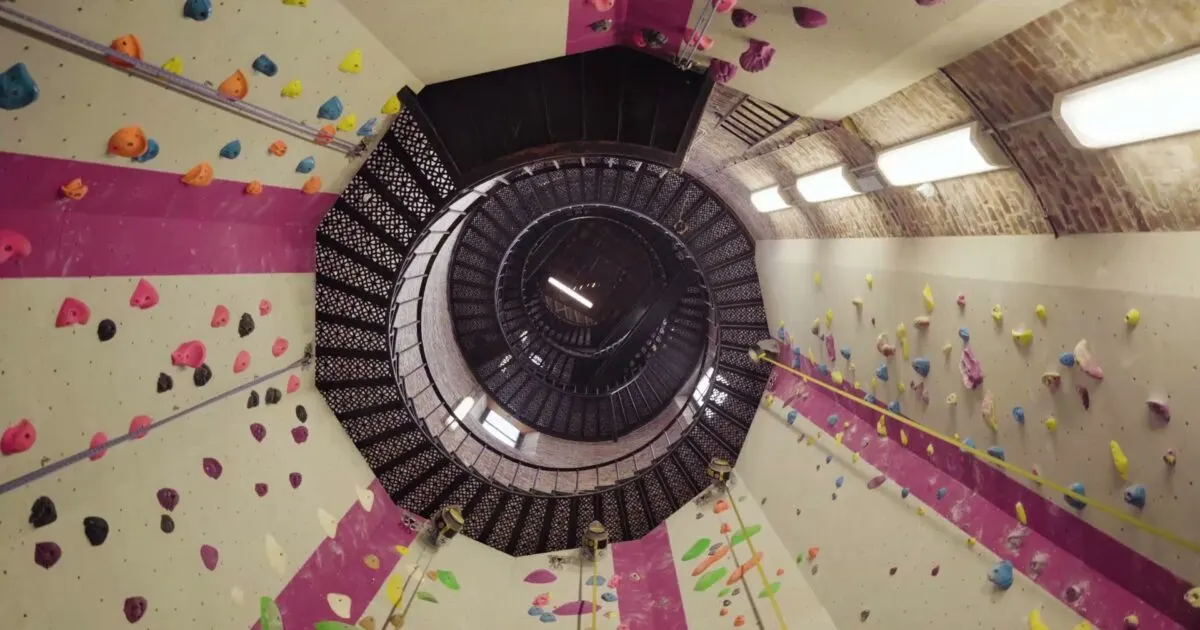 Indoor climbing wall with colorful holds arranged around a central spiral staircase in an urban climbing gym.