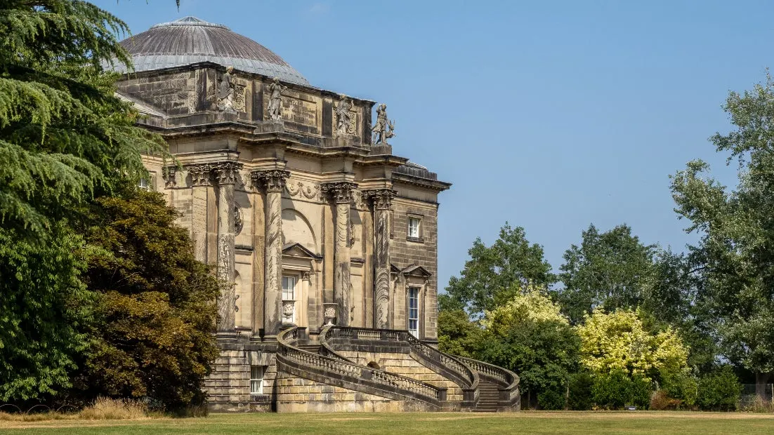 A grand stone neoclassical building with a domed roof and ornamental columns stands in parkland surrounded by mature trees.