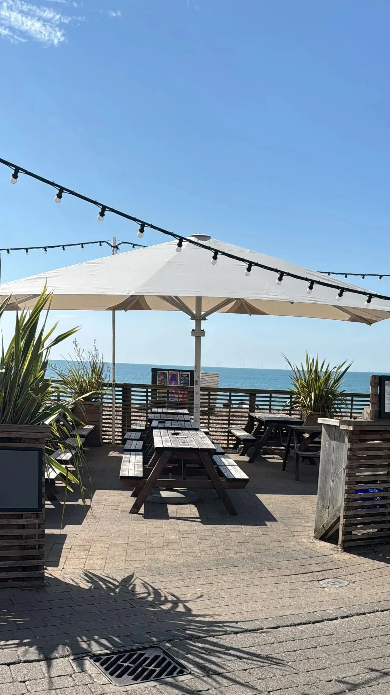 Beachfront terrace with picnic tables sheltered by a large white canopy, sea view beyond wooden railings.