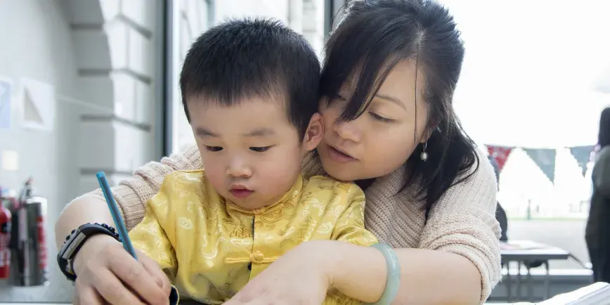 Woman helping young child in yellow jacket hold a paintbrush during an art activity indoors.