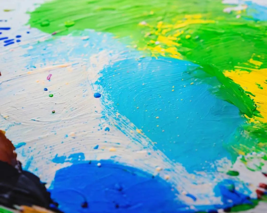 Child's hand painting with bright blue, green, and yellow paint on white paper.