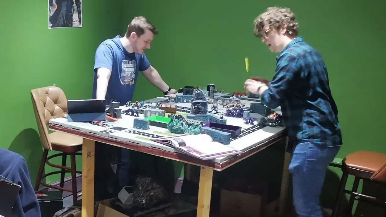 Two players facing each other across a table playing a tabletop wargame with painted miniature figures and scenery.