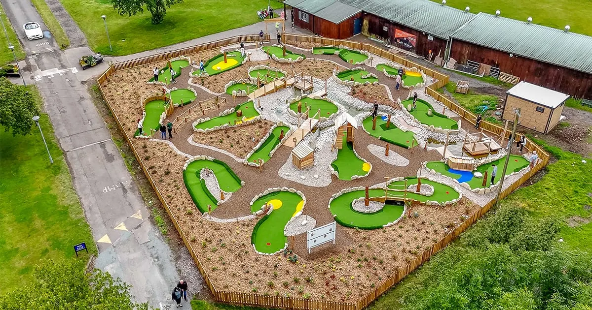 Aerial view of a mini golf course with multiple bright green holes, wooden obstacles, and players scattered across the enclosed sand and gravel layout.